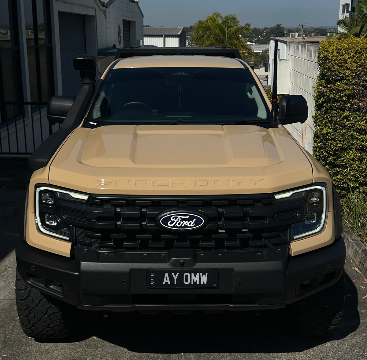 A tan Ford Ranger Super Duty off-road vehicle parked in a driveway outdoors, facing the camera. The truck features a black front grille with the Ford logo, a black bumper, and a roof rack.