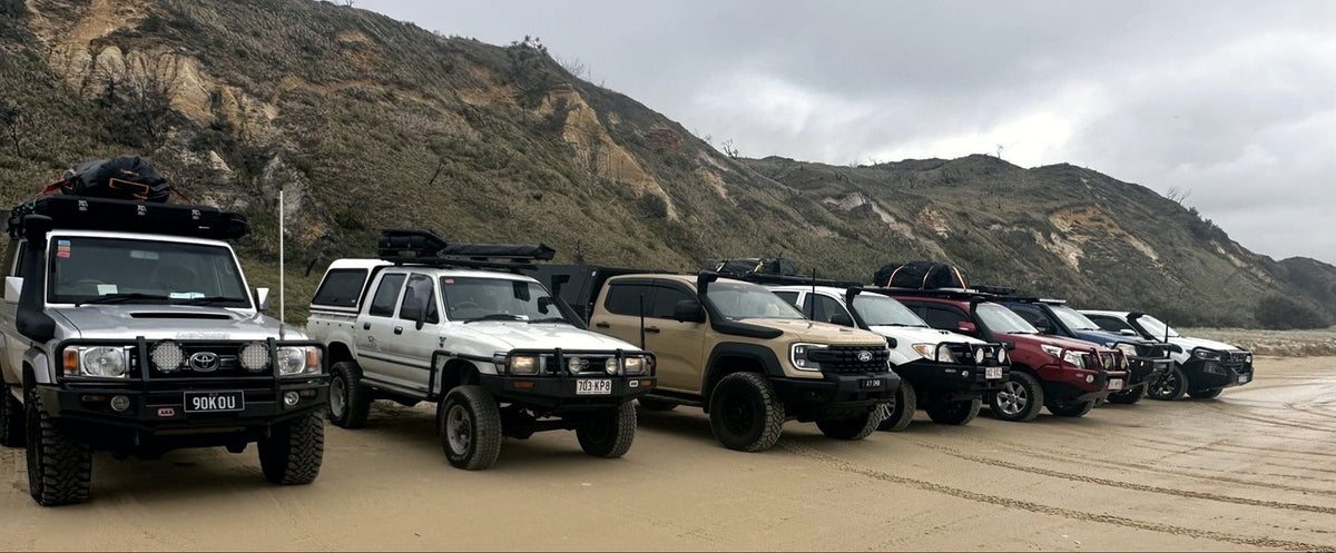 A lineup of off-road SUVs parked on a sandy beach with rocky hills in the background under an overcast sky.